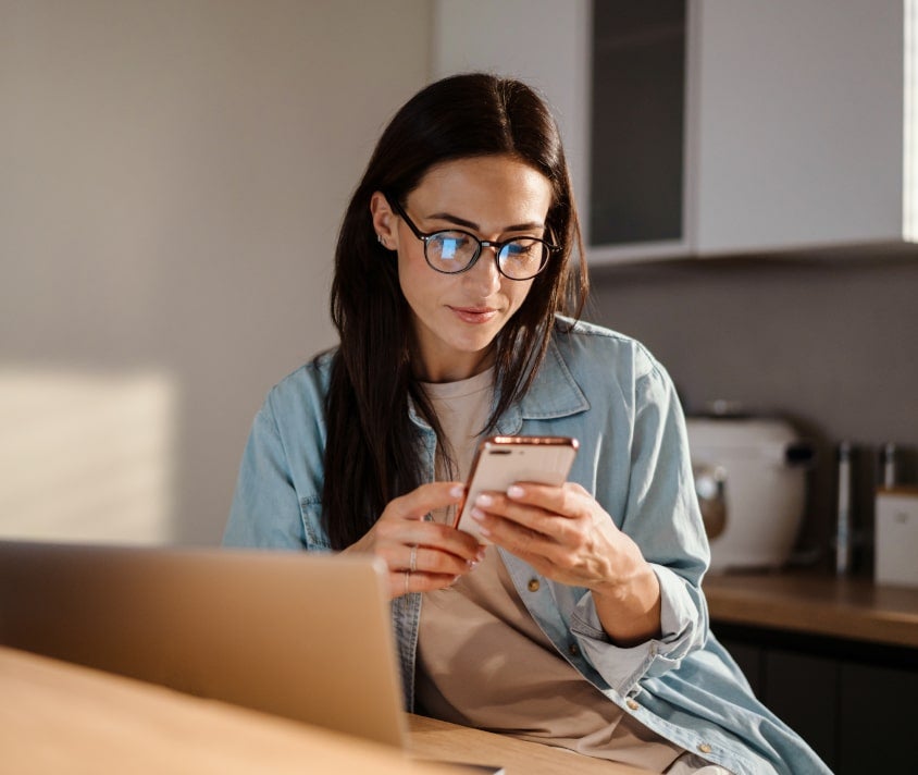 Žena s okuliarmi si prezerá smartfón vo svetlom interiéri kuchyne.