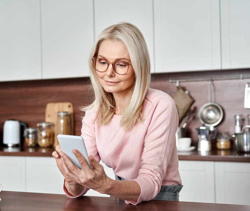 Staršia žena s okuliarmi si v kuchyni prezerá obsah svojho smartfónu.